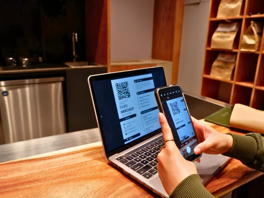 A person scans a digital resume displayed on a laptop using their smartphone in a modern workspace