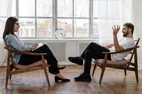 A woman taking notes during a one-on-one conversation with a man in a bright, modern room, illustrating mentorship, coaching, or reflective discussion.