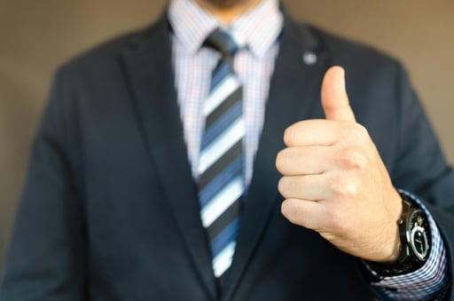 Close-up of a man in a suit giving a thumbs-up gesture, symbolizing approval, success, or positive feedback in a professional context.