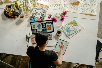 Person engaging in creative design work using a tablet and laptop, surrounded by artistic materials—illustrating active learning through hands-on experience.