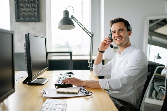 A smiling professional wearing a headset at a desk, ready to communicate